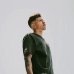 A young man with tattoos stands confidently outdoors on a sunny day in Medellín.