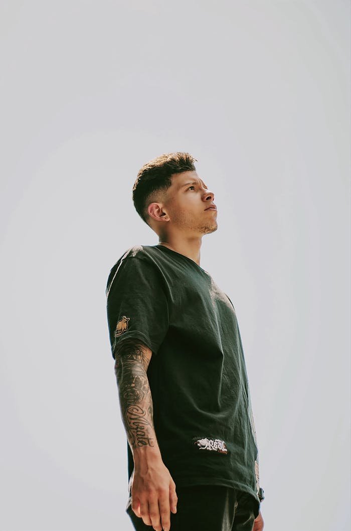 A young man with tattoos stands confidently outdoors on a sunny day in Medellín.