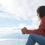 A woman in casual attire enjoys a hot drink while gazing at the sea, epitomizing relaxation.