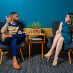 Two professionals in business attire engage in a thoughtful discussion sitting in a stylish, modern room.