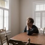 A man sits alone at a table in a bright room, displaying deep contemplation.