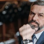 A thoughtful, bearded man in a suit poses indoors, exuding style and confidence.