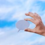 Hand holding a speech bubble against a vibrant blue sky with scattered clouds.